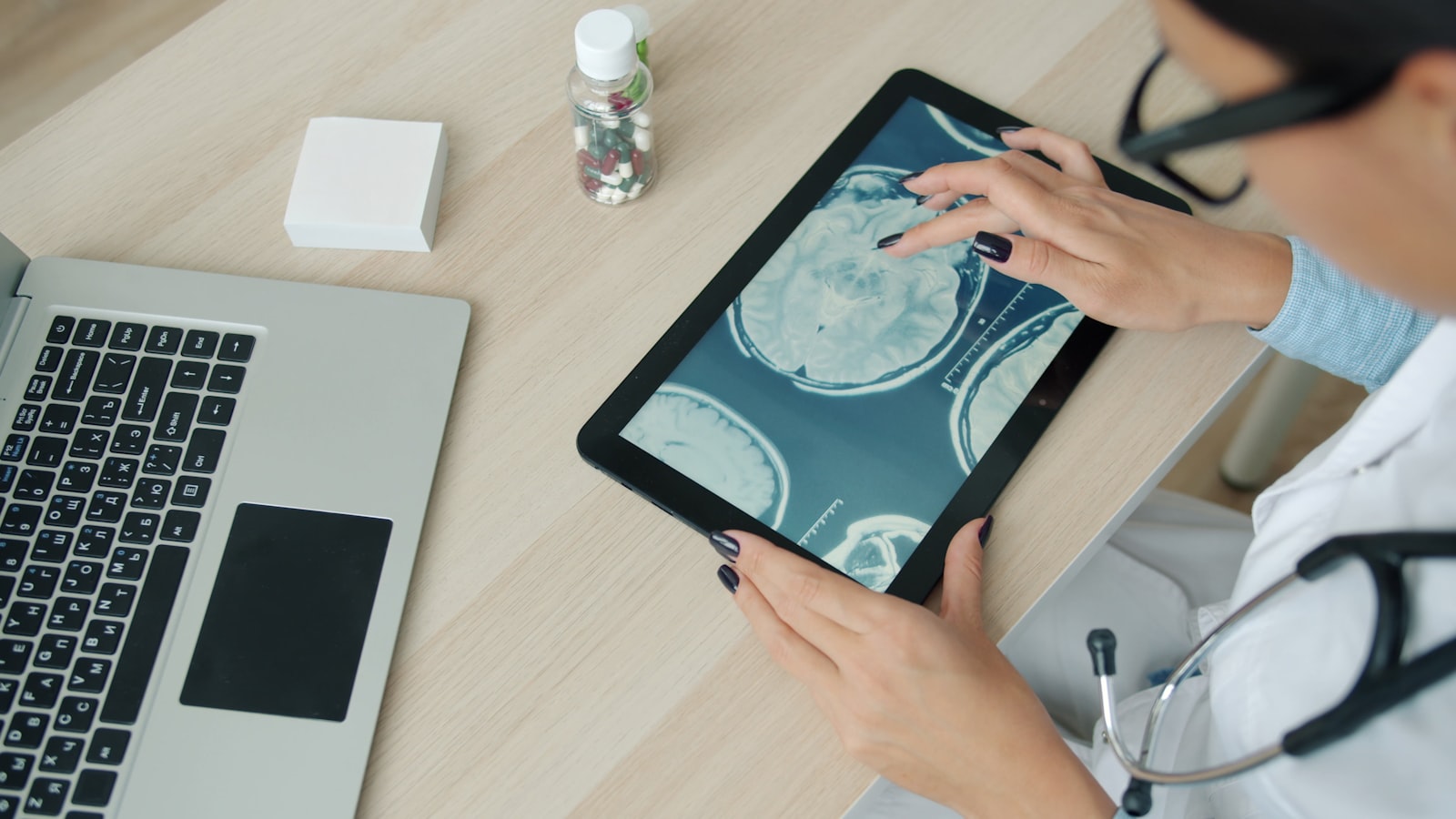 A doctor's hands holding a tablet displaying a brain scan at a desk, with a laptop and pill bottle nearby.