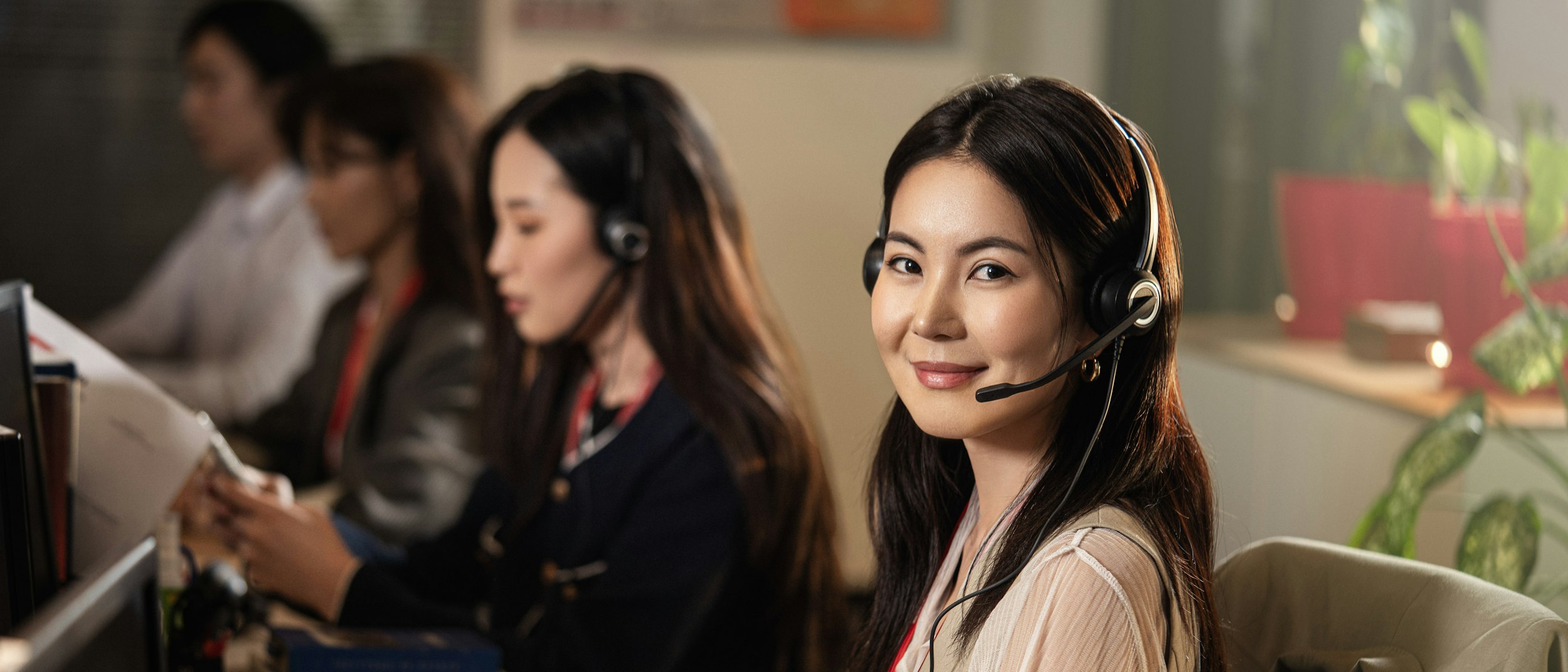 A customer service agent wearing a headset and smiling at the camera, with colleagues wearing headsets working at desks behind her in an office.