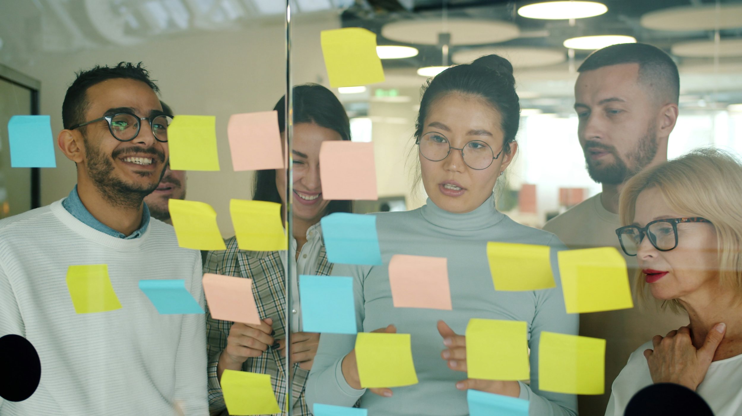 A team of colleagues smiling while arranging colorful sticky notes on a glass wall.