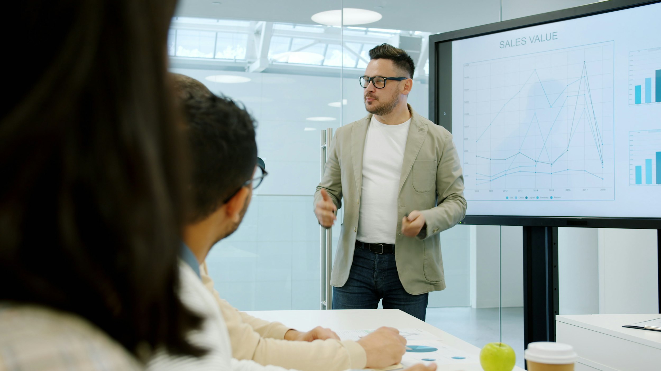 A presenter gesturing toward a large screen showing a sales-value line chart in a meeting room.