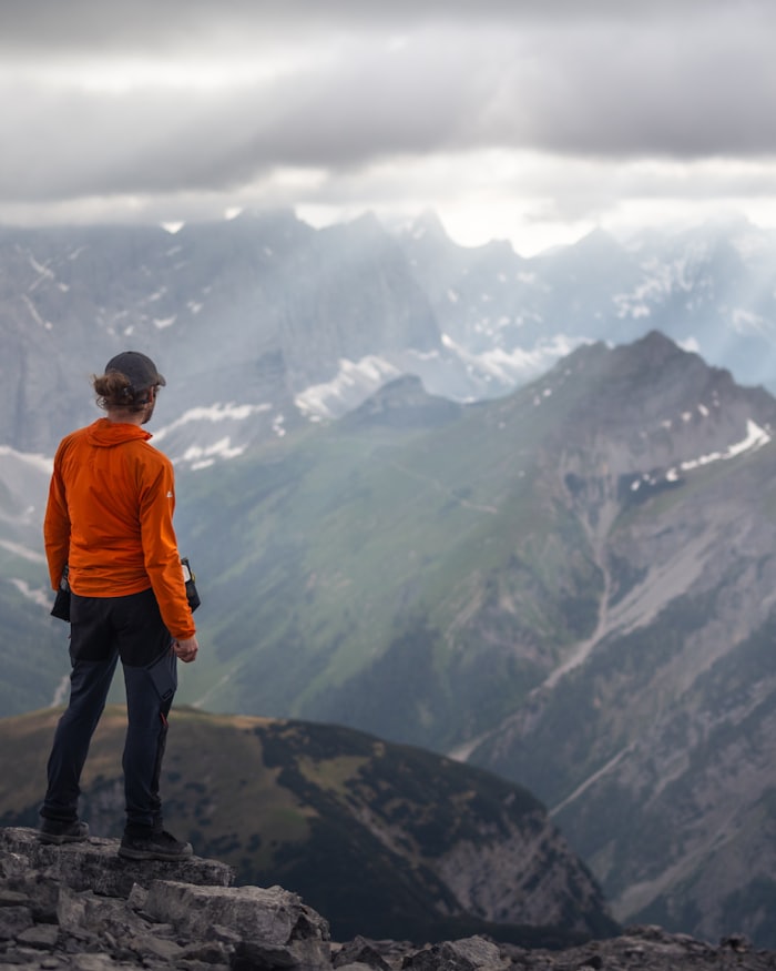 A hiker in an orange jacket standing on a rocky mountain summit looking out over a distant mountain range.