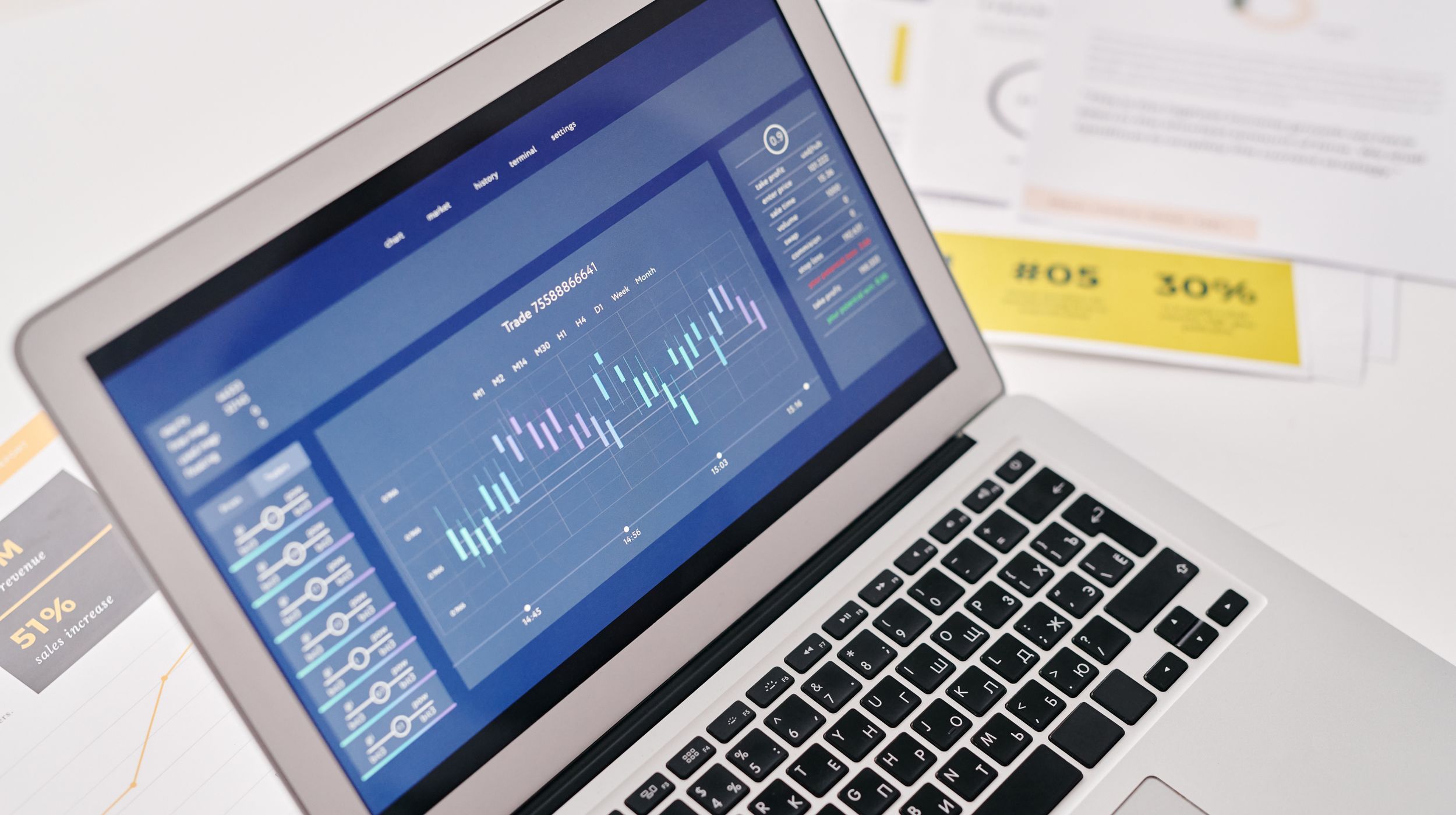An open laptop displaying a financial trading chart, resting on a desk beside printed reports.