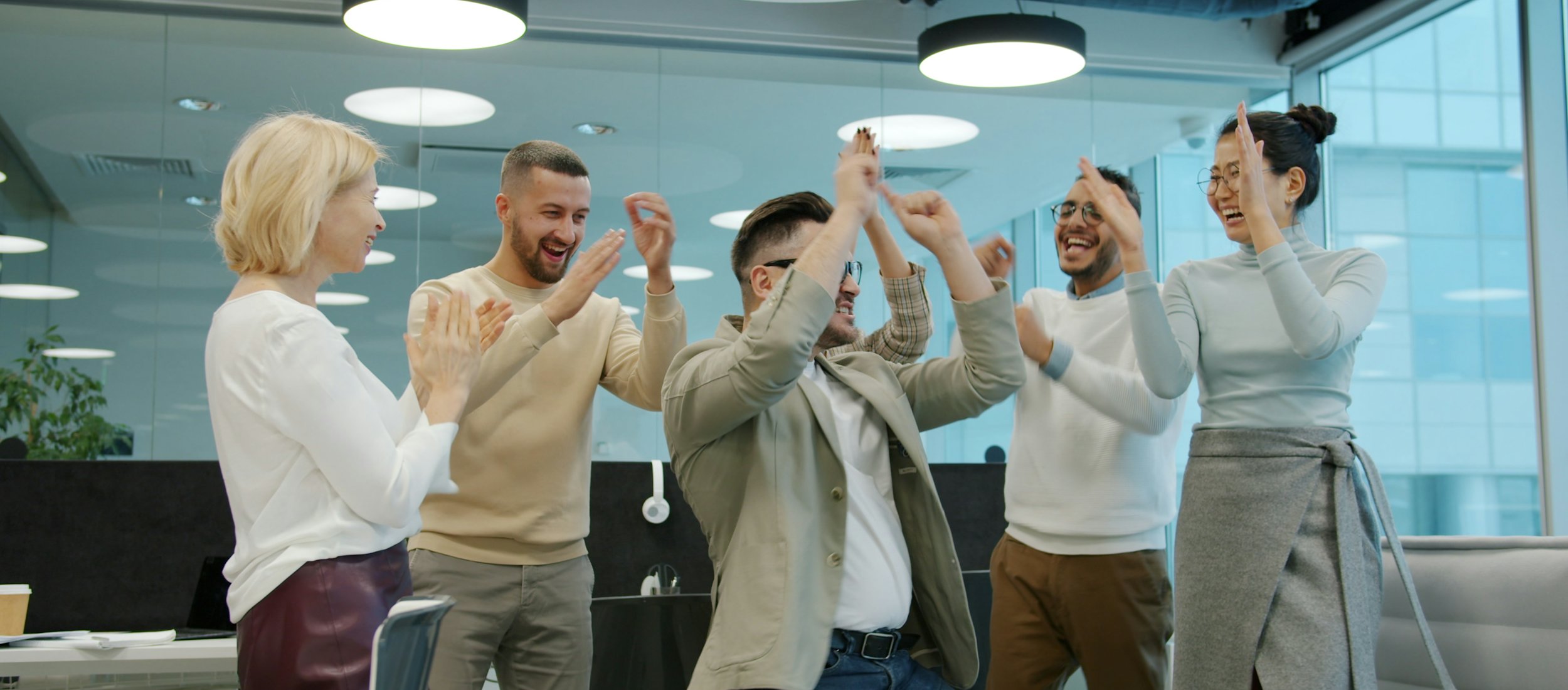 A group of colleagues in a bright glass-walled office raising their hands together in a high-five.