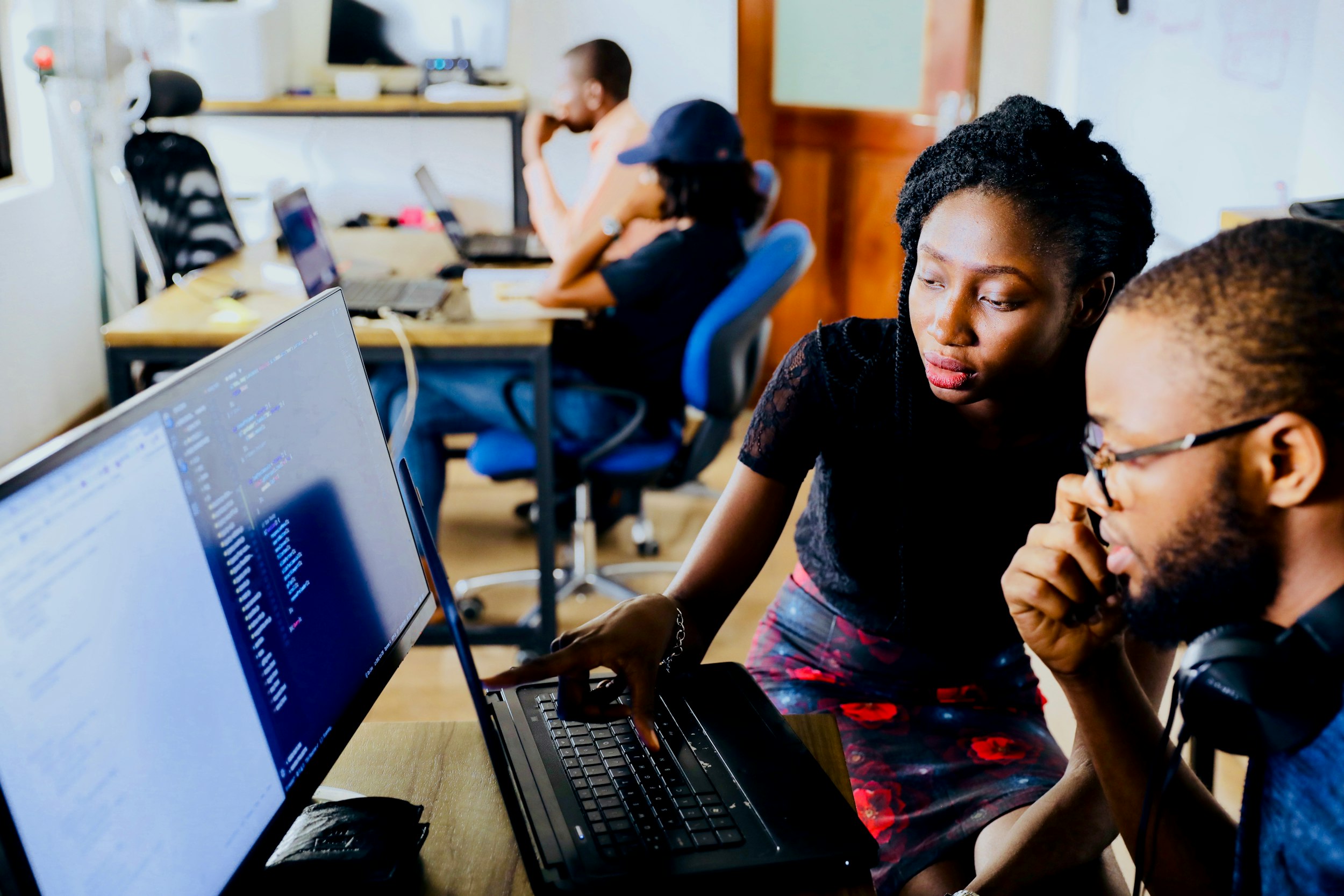 Two colleagues reviewing code on a laptop and an external monitor in an open-plan office, with coworkers at desks in the background.