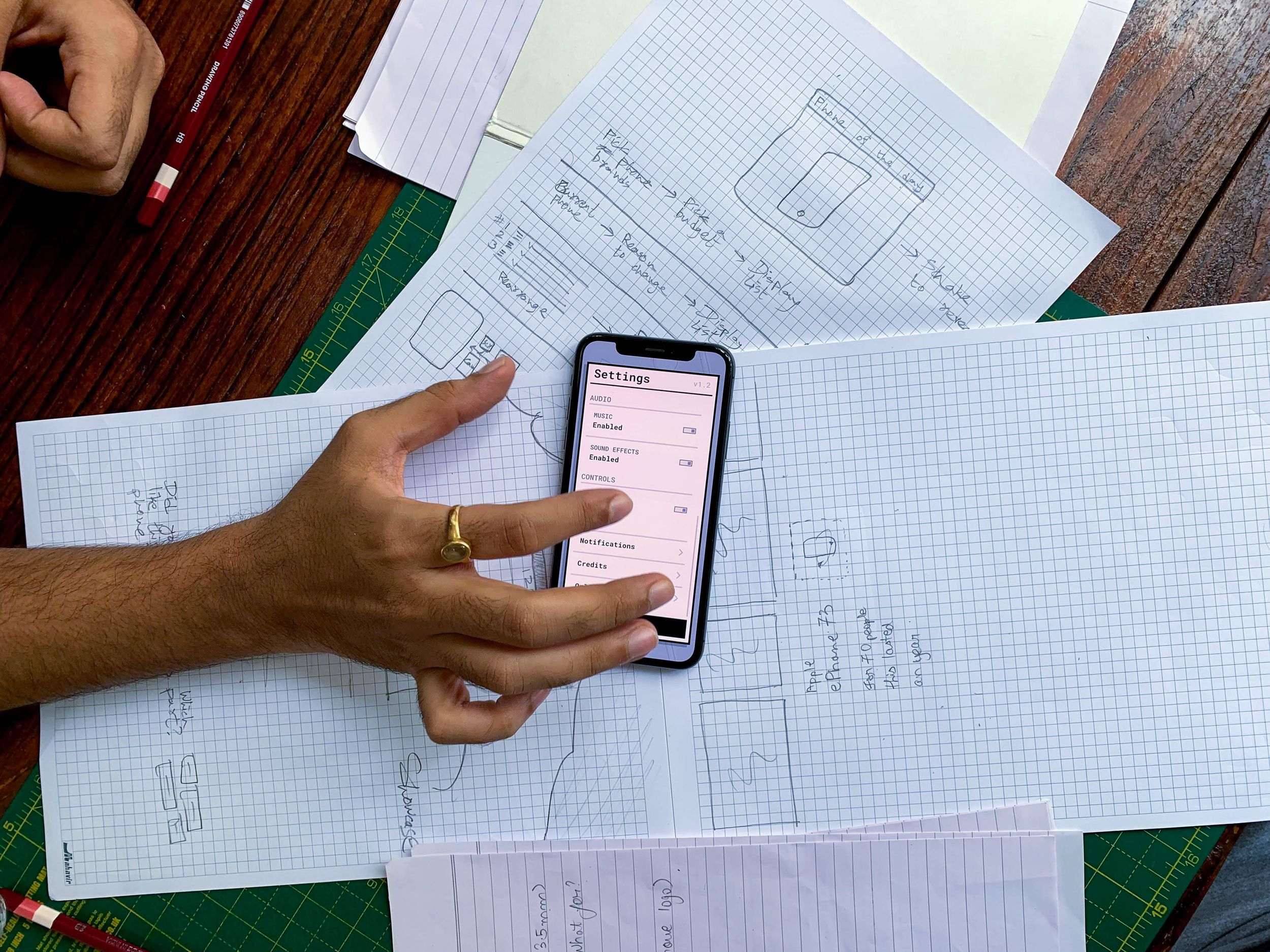 A hand resting a smartphone that displays an app settings screen on top of hand-drawn interface wireframes on graph paper, on a wooden desk with a green cutting mat.